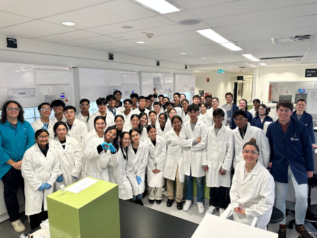 A few dozen students in white labcoats standing in a clean, brightly lit lab. On each side of the picture there is one person in a blue labcoat.